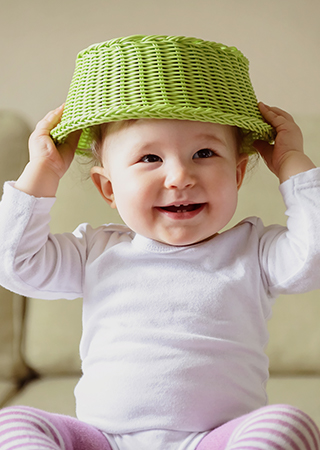 Smiling baby holding green woven basket over head.