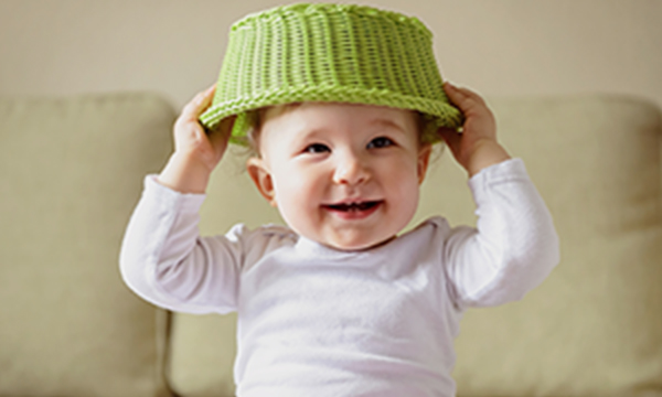 Smiling baby holding green woven basket over head.